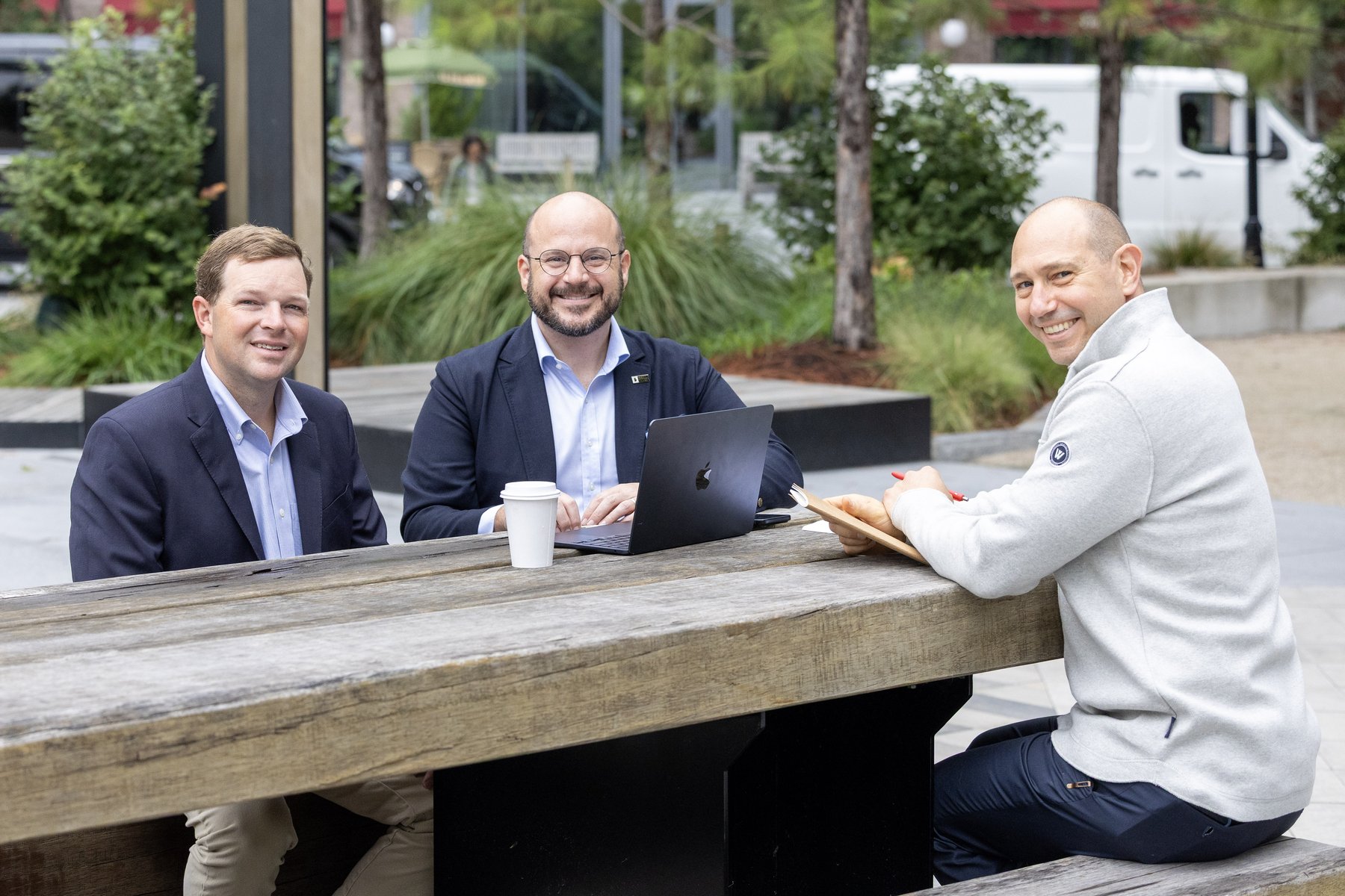 Three men smiling at an outdoor wooden table with a laptop and coffee